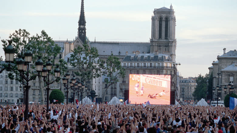 Una pantalla grande que muestra un partido de fútbol a la multitud. Edificios europeos al fondo.